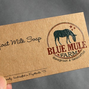 May include: A rectangular, tan-colored card with the words "Goat Milk Soap" in black script. The card features a circular logo with a blue mule silhouette and the text "BLUE MULE FARM" in red. The text "Homegrown & Handcrafted" is also present.