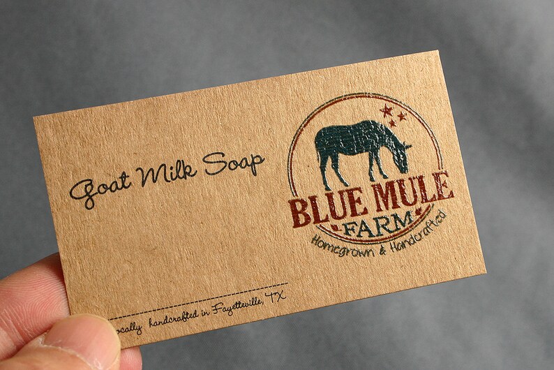 May include: A rectangular, tan-colored card with the words "Goat Milk Soap" in black script. The card features a circular logo with a blue mule silhouette and the text "BLUE MULE FARM" in red. The text "Homegrown & Handcrafted" is also present.