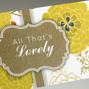 May include: A white card with a brown label that says "All That's Lovely" in white text. The card has a floral pattern with yellow flowers and green leaves.