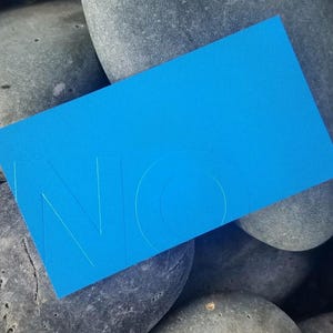 May include: A rectangular bright blue card featuring the embossed letters NO in a clean sans-serif font. The card has a smooth matte finish and is positioned at an angle, displaying a bold monochromatic color scheme with subtle depth.