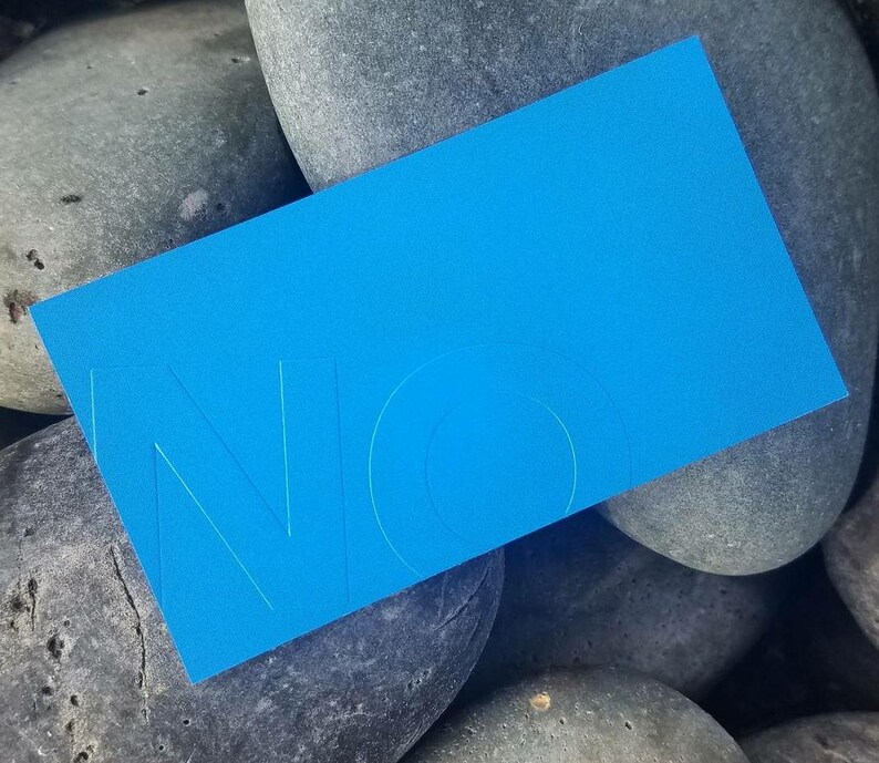 May include: A rectangular bright blue card featuring the embossed letters NO in a clean sans-serif font. The card has a smooth matte finish and is positioned at an angle, displaying a bold monochromatic color scheme with subtle depth.
