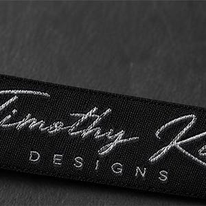 May include: A rectangular black fabric clothing label featuring the embroidered white text Timothy King in a cursive script font, with the word DESIGNS printed in a simple sans-serif font directly underneath the name.