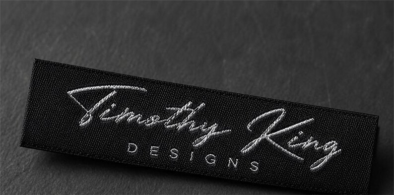 May include: A rectangular black fabric clothing label featuring the embroidered white text Timothy King in a cursive script font, with the word DESIGNS printed in a simple sans-serif font directly underneath the name.