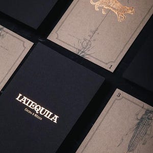 May include: Black rectangular menus with the gold text LATEQUILA COCINA D MEXICO and tan cards featuring gold foil illustrations of a jaguar and a rabbit, along with botanical drawings of corn and agave plants, labelled with the numbers 1 and 3.
