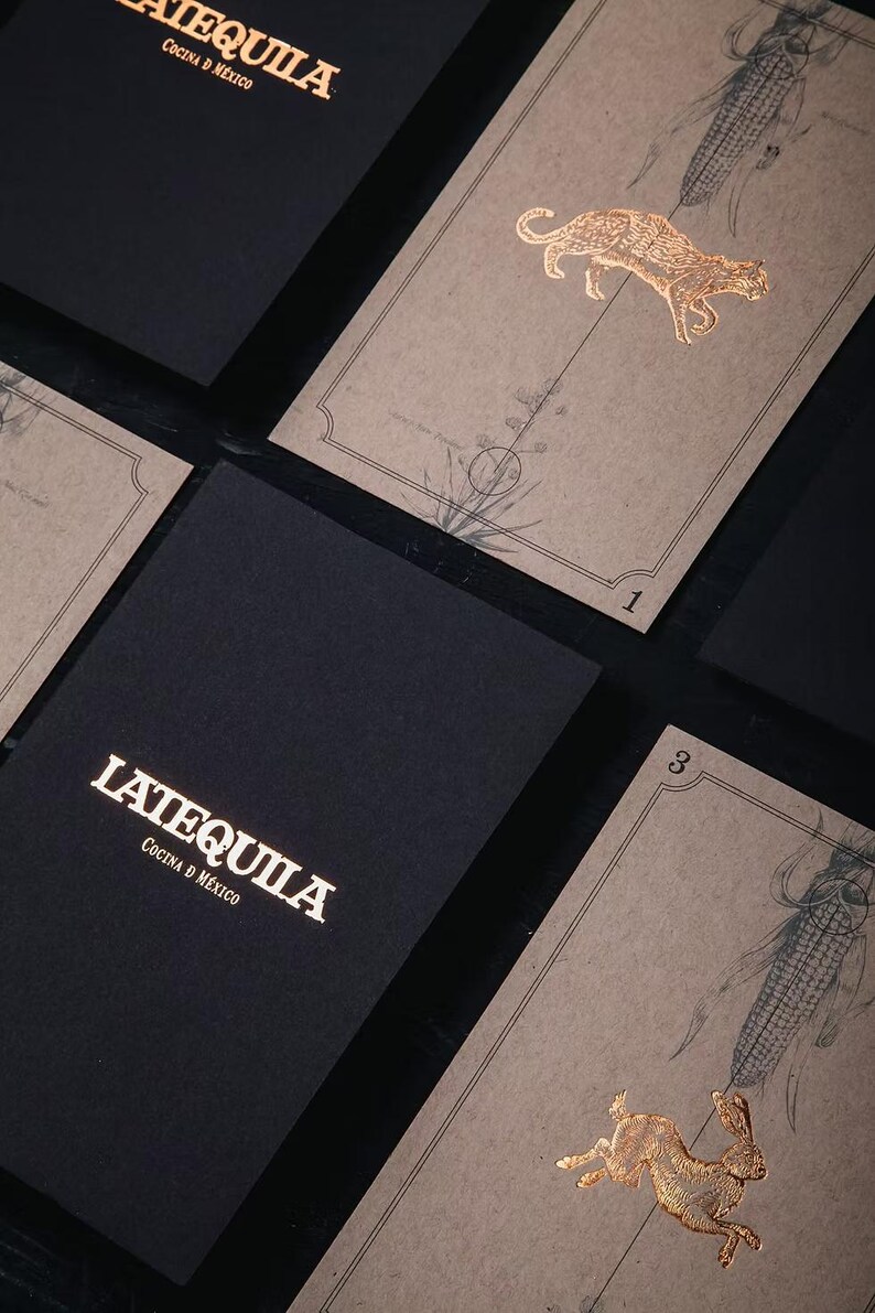 May include: Black rectangular menus with the gold text LATEQUILA COCINA D MEXICO and tan cards featuring gold foil illustrations of a jaguar and a rabbit, along with botanical drawings of corn and agave plants, labelled with the numbers 1 and 3.