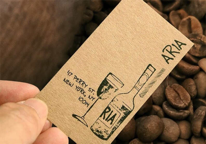 May include: A rectangular tan card featuring a black ink stamp of a wine bottle and glass. The text reads ARIA, 117 PERRY ST. NEW YORK, NY 10014. The card has a textured, fibrous paper appearance with a simple, hand-drawn style graphic illustration.