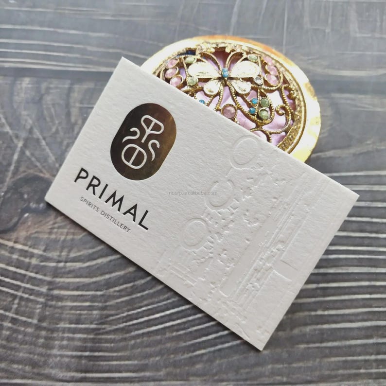 May include: A rectangular white business card with a gold foil logo and the text PRIMAL SPIRITS DISTILLERY, featuring an embossed architectural pattern. Behind the card sits a round gold metal compact with a filigree lid and pink inlay.