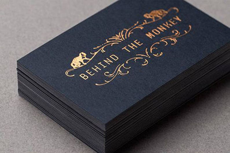 May include: A stack of rectangular business cards made of dark navy blue cardstock. The top card features gold foil stamping with the text BEHIND THE MONKEY, accompanied by decorative filigree flourishes and small monkey illustrations on each side.