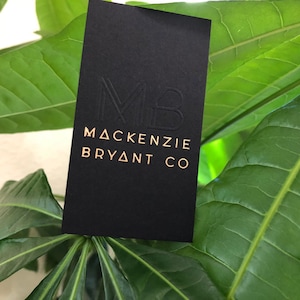 May include: A rectangular black business card featuring a debossed MB monogram at the top and the gold-colored text MACKENZIE BRYANT CO printed below. The card has a smooth matte finish and is positioned vertically against a background of green leaves.