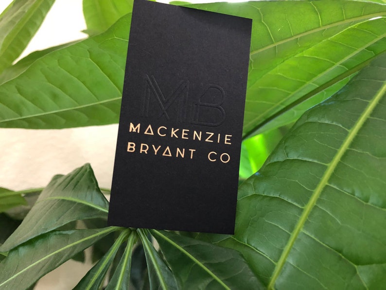May include: A rectangular black business card featuring a debossed MB monogram at the top and the gold-colored text MACKENZIE BRYANT CO printed below. The card has a smooth matte finish and is positioned vertically against a background of green leaves.