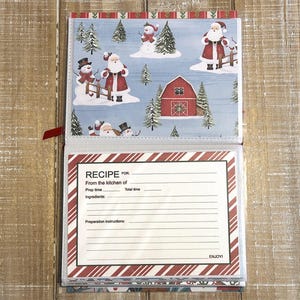 Puede incluir: Libro de recetas abierto con tem&aacute;tica navide&ntilde;a. La p&aacute;gina superior presenta una escena invernal con mu&ntilde;ecos de nieve, Pap&aacute; Noel y un granero rojo. La p&aacute;gina inferior tiene una plantilla de receta con espacio para ingredientes e instrucciones. El libro est&aacute; encuadernado con una cinta roja.