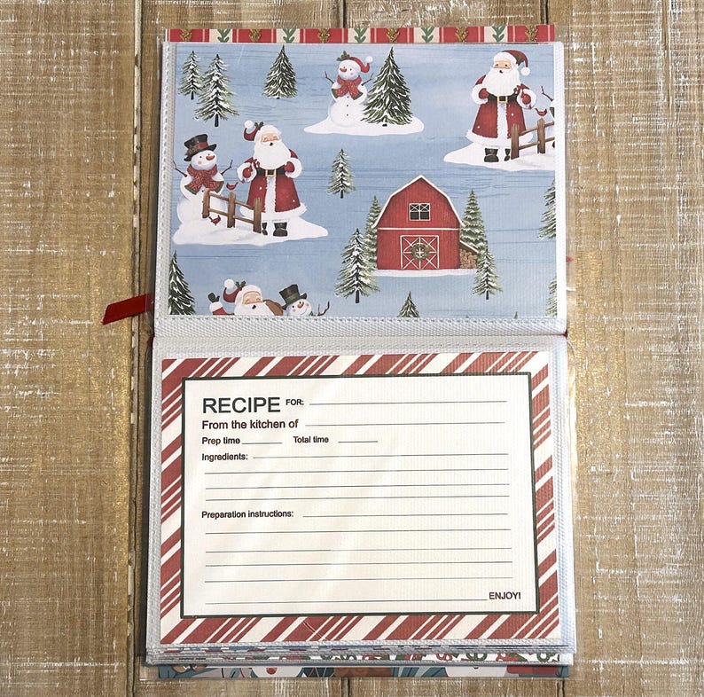 Puede incluir: Libro de recetas abierto con tem&aacute;tica navide&ntilde;a. La p&aacute;gina superior presenta una escena invernal con mu&ntilde;ecos de nieve, Pap&aacute; Noel y un granero rojo. La p&aacute;gina inferior tiene una plantilla de receta con espacio para ingredientes e instrucciones. El libro est&aacute; encuadernado con una cinta roja.
