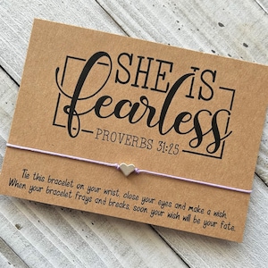May include: A brown card with the text "She is fearless - Proverbs 31:25" printed on it. A purple string bracelet with a silver heart charm is attached to the card. The card also includes instructions for making a wish when the bracelet breaks.