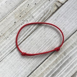 Puede incluir: Un brazalete de cuerda roja con un cierre simple de nudo. El brazalete est&aacute; sobre un fondo de madera blanca.