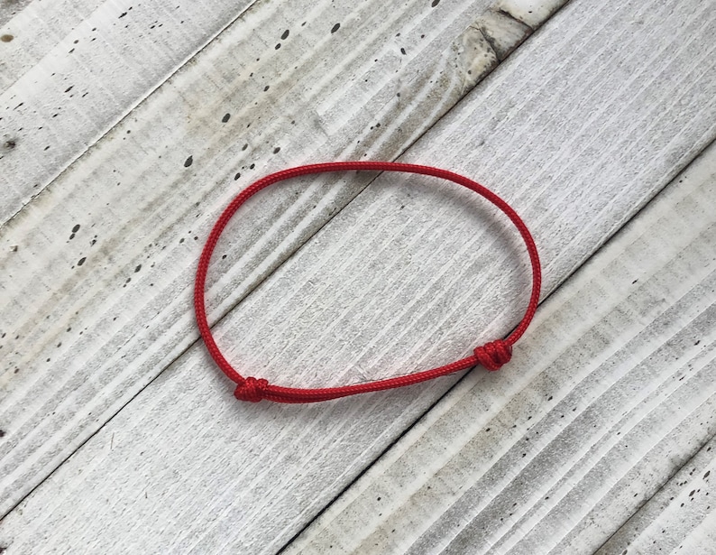 Puede incluir: Un brazalete de cuerda roja con un cierre simple de nudo. El brazalete est&aacute; sobre un fondo de madera blanca.