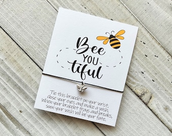 May include: A white card with the words "Bee You tiful" and a small bee illustration. A black string bracelet with a bee charm is attached. The card includes text about making a wish.