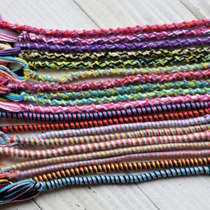 May include: A collection of colorful friendship bracelets made with various types of string and yarn. The bracelets are woven in different patterns and colors, including pink, purple, blue, green, yellow, and black.