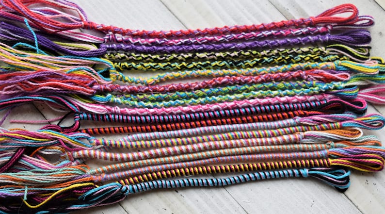 May include: A collection of colorful friendship bracelets made with various types of string and yarn. The bracelets are woven in different patterns and colors, including pink, purple, blue, green, yellow, and black.