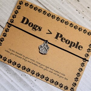 May include: A brown card with black paw prints around the border. The card has the text "Dogs > People" and a black string with a silver paw print charm. The card also has instructions for making a wish.