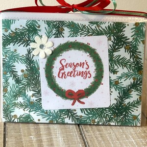 Puede incluir: Una caja de regalo envuelta en celof&aacute;n transparente, atada con una cinta roja y verde. La caja presenta un dise&ntilde;o navide&ntilde;o con una corona y las palabras "Season's Greetings". Un adorno floral blanco est&aacute; adjunto.