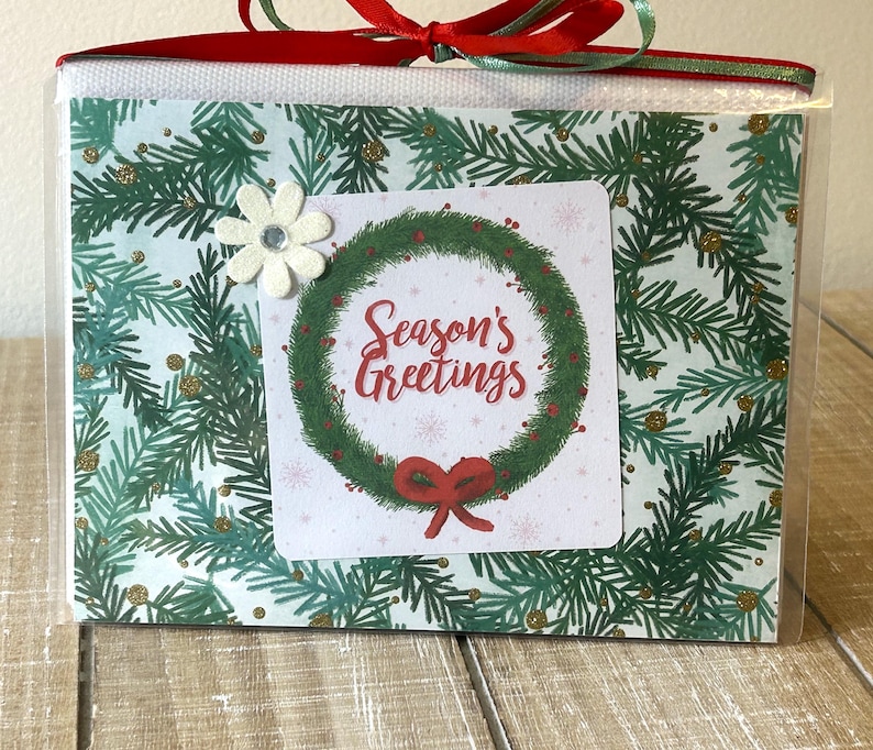 Puede incluir: Una caja de regalo envuelta en celof&aacute;n transparente, atada con una cinta roja y verde. La caja presenta un dise&ntilde;o navide&ntilde;o con una corona y las palabras "Season's Greetings". Un adorno floral blanco est&aacute; adjunto.
