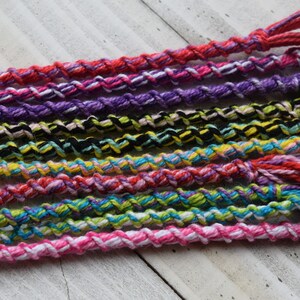 May include: A collection of colorful friendship bracelets made with braided string. The bracelets are in various colors, including pink, purple, red, yellow, blue, green, and black.