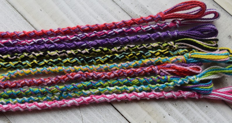 May include: A collection of colorful friendship bracelets made with braided string. The bracelets are in various colors, including pink, purple, red, yellow, blue, green, and black.