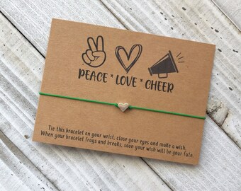 May include: A green string bracelet with a silver heart charm. The card attached to the bracelet reads "Peace * Love * Cheer" and includes instructions to tie the bracelet on your wrist, close your eyes, make a wish, and when the bracelet frays, your wish will come true.