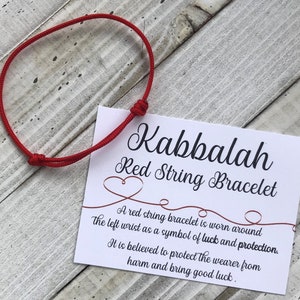 Puede incluir: Pulsera de cuerda roja con una tarjeta blanca que dice "Kabbalah Red String Bracelet". Una pulsera de cuerda roja se usa alrededor de la mu&ntilde;eca izquierda como s&iacute;mbolo de suerte y protecci&oacute;n. Se cree que protege al usuario del da&ntilde;o y trae buena suerte.