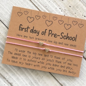 Puede incluir: Una tarjeta con el texto "first day of Pre-School" y un mensaje sobre dos pulseras, una grande y una pequeña. El mensaje dice "Llevaré la grande y pensaré en ti. La pequeña es para ti, así que piensa en mí también. Llévala a la escuela mientras aprendes y juegas, Para tener mi corazón contigo mientras estás lejos."