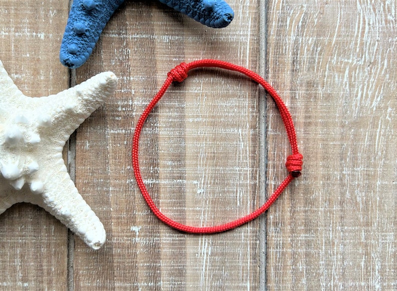 Puede incluir: Un brazalete de cuerda roja con un nudo en cada extremo. El brazalete est&aacute; sobre una superficie de madera.