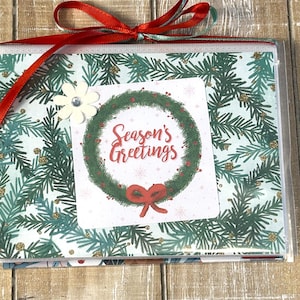 Puede incluir: Un juego de tarjetas de Navidad envueltas en celof&aacute;n transparente, atadas con una cinta roja y un adorno floral blanco. Las tarjetas presentan una corona verde con la frase "Season's Greetings" y un patr&oacute;n de ramitas.