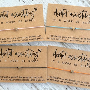 Peut inclure: Quatre cartes brunes avec le texte "dental assisting is a work of heart" et un petit charme de c&oelig;ur en or attach&eacute; &agrave; un bracelet en ficelle. Chaque carte a un bracelet en ficelle de couleur diff&eacute;rente&nbsp;: rose, bleu, noir et orange.