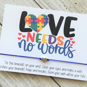 Op de afbeelding: Een witte kaart met de tekst "LOVE NEEDS NO WORDS" in regenboogkleuren. Een puzzelhart bevindt zich boven de tekst. Een blauw koordarmband met een zilveren hartbedel is aan de kaart bevestigd. De kaart luidt "Bind deze armband om je pols Sluit je ogen en wens iets Als je armband verslijt en breekt, zal je wens snel je lot zijn."