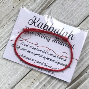 Puede incluir: Pulsera de cuerda roja con una tarjeta blanca que dice "Kabbalah Red String Bracelet". Una pulsera de cuerda roja se usa alrededor de la mu&ntilde;eca izquierda como s&iacute;mbolo de suerte y protecci&oacute;n. Se cree que protege al usuario del da&ntilde;o y trae buena suerte.