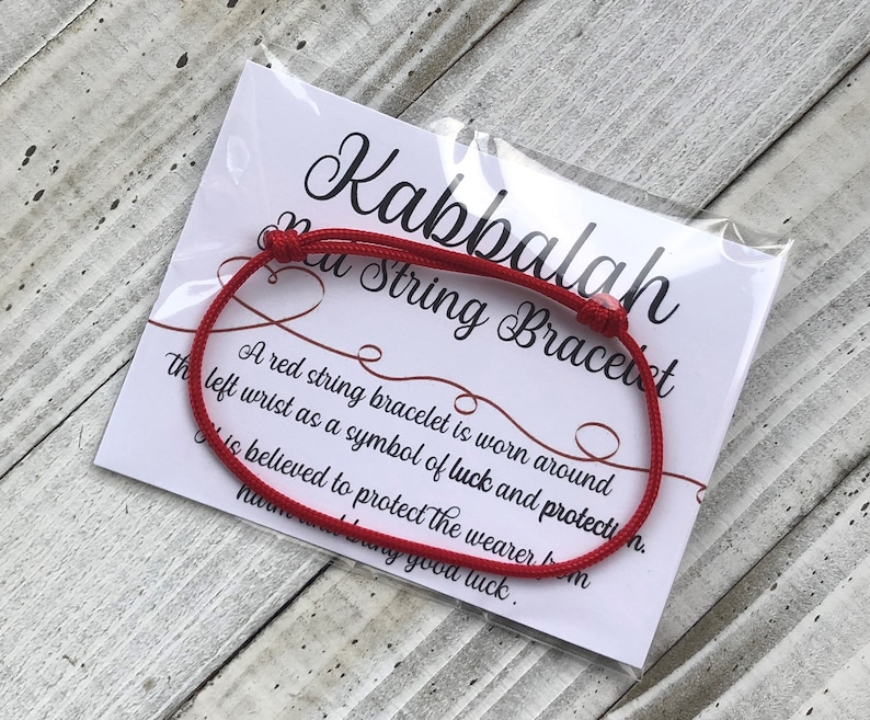 Puede incluir: Pulsera de cuerda roja con una tarjeta blanca que dice "Kabbalah Red String Bracelet". Una pulsera de cuerda roja se usa alrededor de la mu&ntilde;eca izquierda como s&iacute;mbolo de suerte y protecci&oacute;n. Se cree que protege al usuario del da&ntilde;o y trae buena suerte.