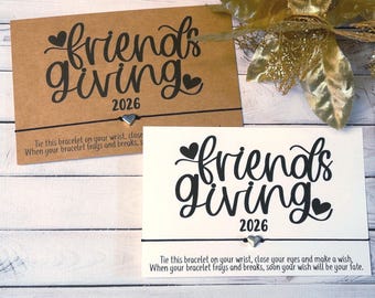May include: Two wish bracelets on rectangular cards, one kraft brown and one white. Each card features the black text "friends giving 2026" with heart accents and a thin black string with a small silver heart charm attached to the center.