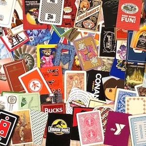 May include: A collection of playing cards and promotional cards with various designs and logos. The cards feature images of cartoon characters, brands, and text, including "Jurassic Park," "Marvel," and "Have Some Fun!"