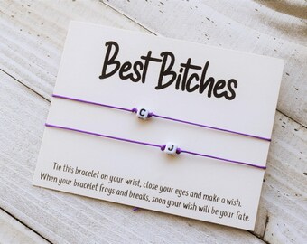Può includere: Una carta bianca con le parole "Best Bitches" in corsivo nero. Sono attaccati due braccialetti di filo viola, ognuno con una perlina con lettere. Il testo sottostante recita: "Allaccia questo braccialetto al tuo polso, chiudi gli occhi ed esprimi un desiderio."