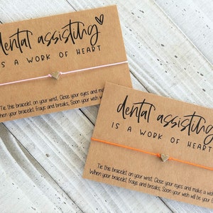 Peut inclure: Deux cartes brunes avec le texte "dental assisting is a work of heart" et un petit charme en forme de c&oelig;ur dor&eacute; attach&eacute; &agrave; un bracelet en ficelle. La ficelle est rose sur une carte et orange sur l'autre.