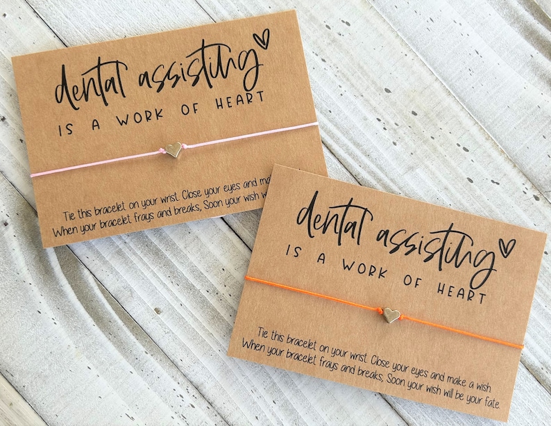 Peut inclure: Deux cartes brunes avec le texte "dental assisting is a work of heart" et un petit charme en forme de c&oelig;ur dor&eacute; attach&eacute; &agrave; un bracelet en ficelle. La ficelle est rose sur une carte et orange sur l'autre.