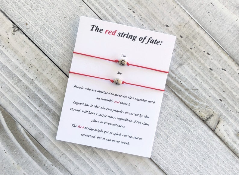 May include: Two red string bracelets with the words "You" and "Me" on them. The bracelets have small silver beads with the letters "C" and "L" on them. The text on the card reads: "The red string of fate: People who are destined to meet are tied together with an invisible red thread. Legend has it that the two people connected by this thread will have a major story, regardless of the time, place or circumstances. The Red String might get tangled, contracted or stretched, but it can never break."