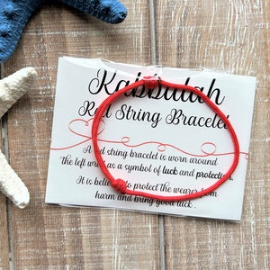 Puede incluir: Pulsera de cuerda roja con una tarjeta blanca que dice "Kabbalah Red String Bracelet". Una pulsera de cuerda roja se usa alrededor de la mu&ntilde;eca izquierda como s&iacute;mbolo de suerte y protecci&oacute;n. Se cree que protege al usuario del da&ntilde;o y trae buena suerte.