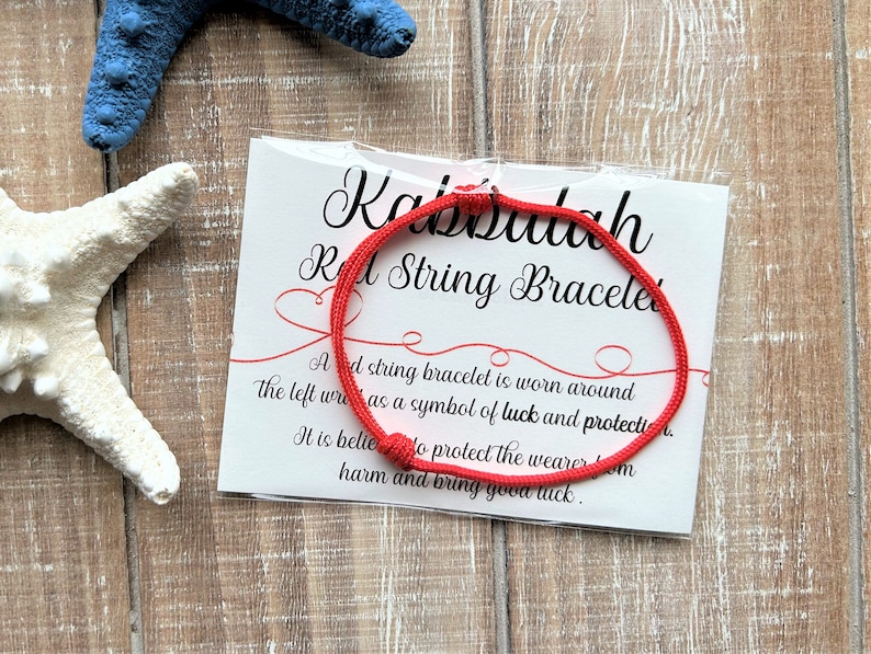 Puede incluir: Pulsera de cuerda roja con una tarjeta blanca que dice "Kabbalah Red String Bracelet". Una pulsera de cuerda roja se usa alrededor de la mu&ntilde;eca izquierda como s&iacute;mbolo de suerte y protecci&oacute;n. Se cree que protege al usuario del da&ntilde;o y trae buena suerte.