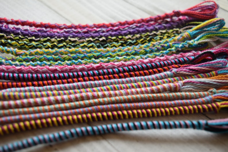 May include: A collection of colorful friendship bracelets made with woven string. The bracelets are in various patterns and colors, including pink, blue, green, yellow, orange, red, black, and white.