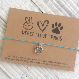 May include: A brown card with the text "Peace Love Paws" and a peace sign, heart, and paw print. A blue string bracelet with a silver paw print charm is attached to the card.