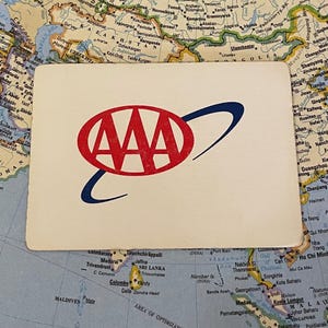 May include: A rectangular card with the AAA logo in red and blue. The logo features three red letter A's and a blue curved line. The card is placed on a world map.