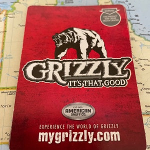 Puede incluir: Tarjeta de producto Grizzly roja y negra con un oso blanco. La tarjeta incluye el texto "GRIZZLY IT'S THAT GOOD" y "mygrizzly.com". También tiene una advertencia sobre el cáncer de boca.