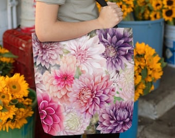 May include: A woman is holding a tote bag with a floral print. The bag is made of canvas and has a black strap. The print features pink, purple, and white dahlias.