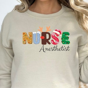 May include: A beige sweatshirt with a Christmas-themed graphic that says "NURSE Anesthetist". The letters are decorated with gingerbread, snowflakes, a reindeer, a Santa hat, and a Christmas tree.